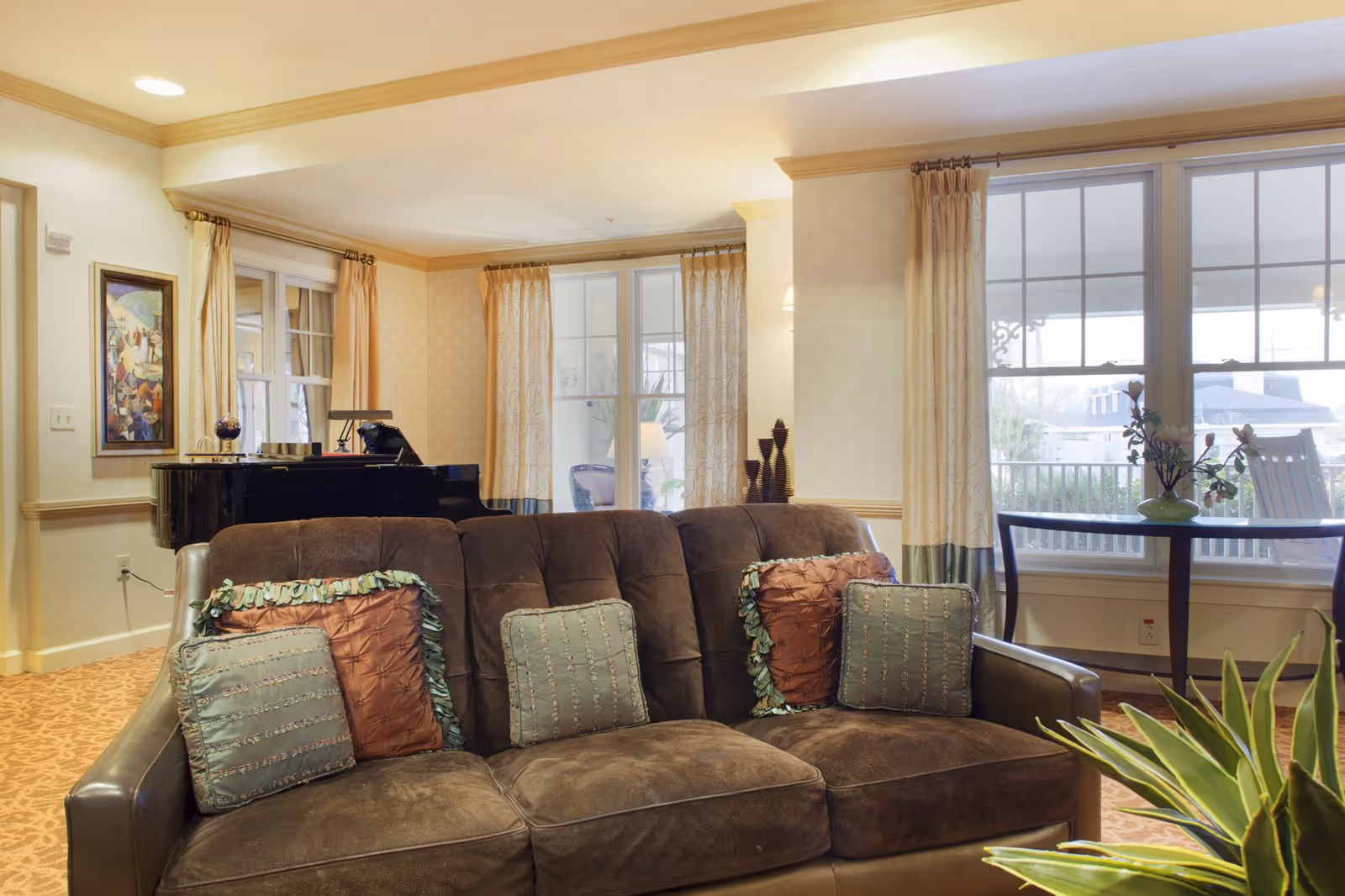 Cozy lounge area with a brown sofa and decorative pillows, a grand piano, large windows, and a table with plants.