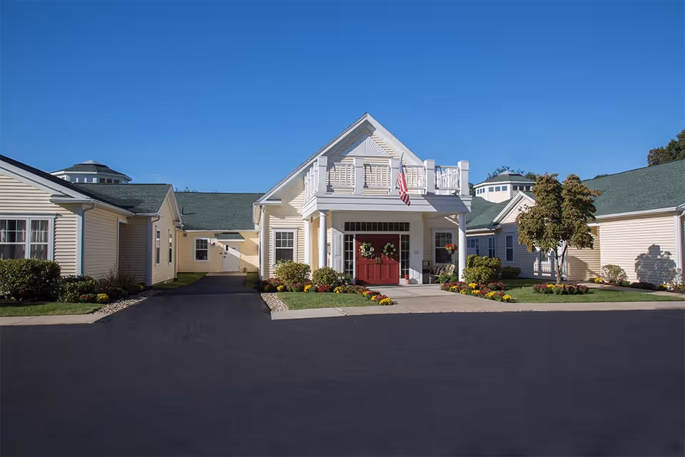 Front entrance of a yellow single-story senior living building with a covered portico, red double doors, an American flag, and landscaped flowerbeds.
