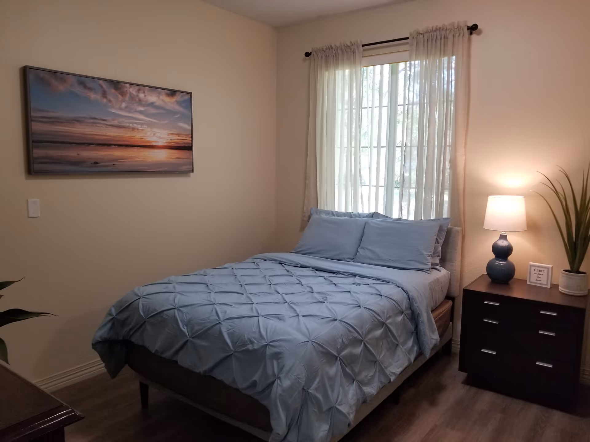 A neatly made bed with light blue bedding and pillows in a bedroom. There is a window with sheer white curtains behind the bed, a dark wooden nightstand with a blue lamp, a small framed sign, and a potted plant. A large framed picture of a sunset over water hangs on the wall to the left.