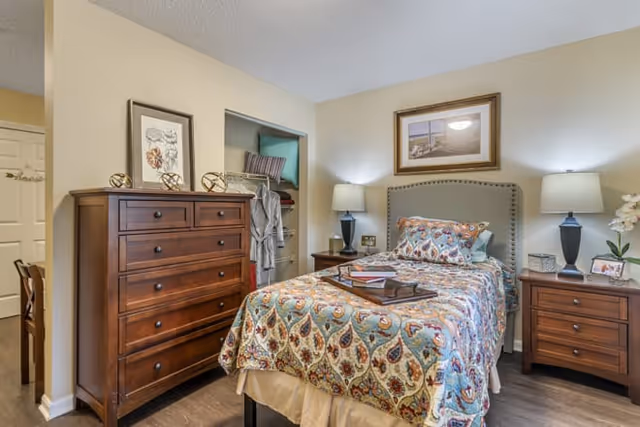 A cozy bedroom featuring a single bed with a patterned bedspread and multiple pillows. On either side of the bed are wooden nightstands with lamps and decorative items. A wooden dresser with drawers is positioned to the left, next to an open closet with hanging clothes. The walls are painted a soft beige, and a framed picture hangs above the bed.