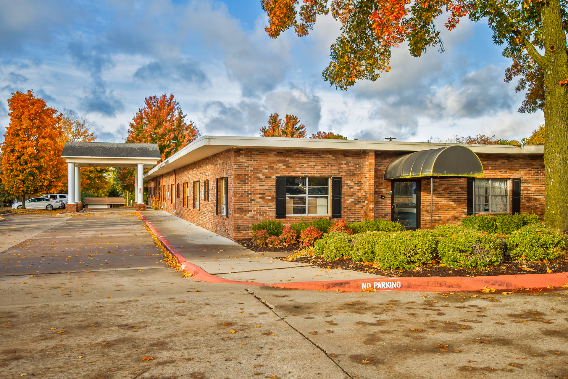 Exterior view of a single-story brick building with black window shutters and a black awning over the entrance. The building is surrounded by green bushes and trees with autumn-colored leaves. A concrete driveway and parking area are visible in front of the building under a partly cloudy sky.