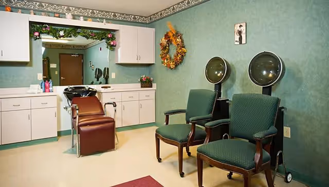 Interior of a salon area in an assisted living facility with two green upholstered chairs under hair dryers, a brown reclining salon chair in front of a sink, white cabinets, a large mirror decorated with flowers, and a fall-themed wreath on the green wall.