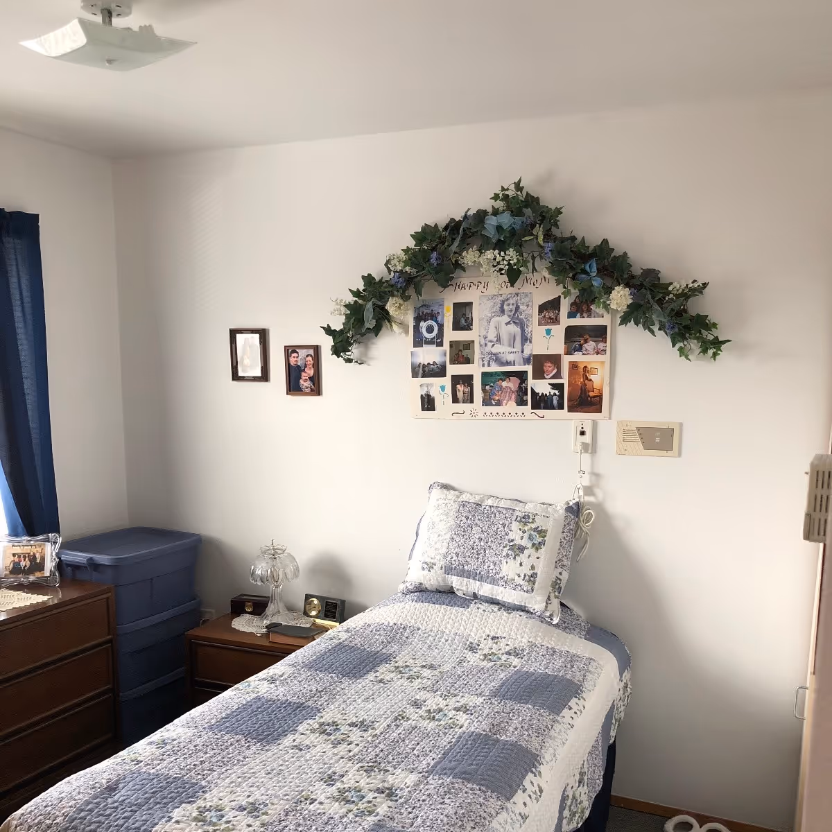 A small bedroom with a single bed covered in a blue and white patchwork quilt and matching pillow. Above the bed is a decorative floral garland and a collage of family photos on the wall. To the left of the bed is a wooden dresser with framed photos and a blue plastic storage bin. A window with dark blue curtains is partially visible on the left side.