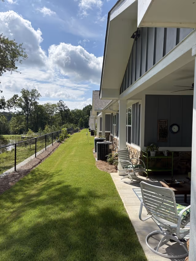 A sunny outdoor view of a grassy area beside a row of residential units with covered patios. The patios have white metal chairs and small tables. A black chain-link fence runs along the left side, separating the grass from a wooded area and a body of water. The sky is partly cloudy with blue patches.