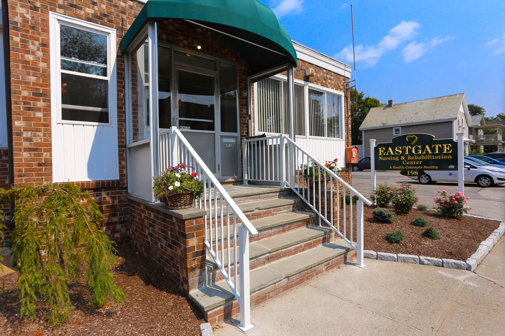 Entrance to Eastgate Nursing & Rehabilitation Center showing brick building with a green awning over the door, white railings along the steps, flower baskets on the ledges, and a sign with the facility name and address near a landscaped area with flowers and shrubs.