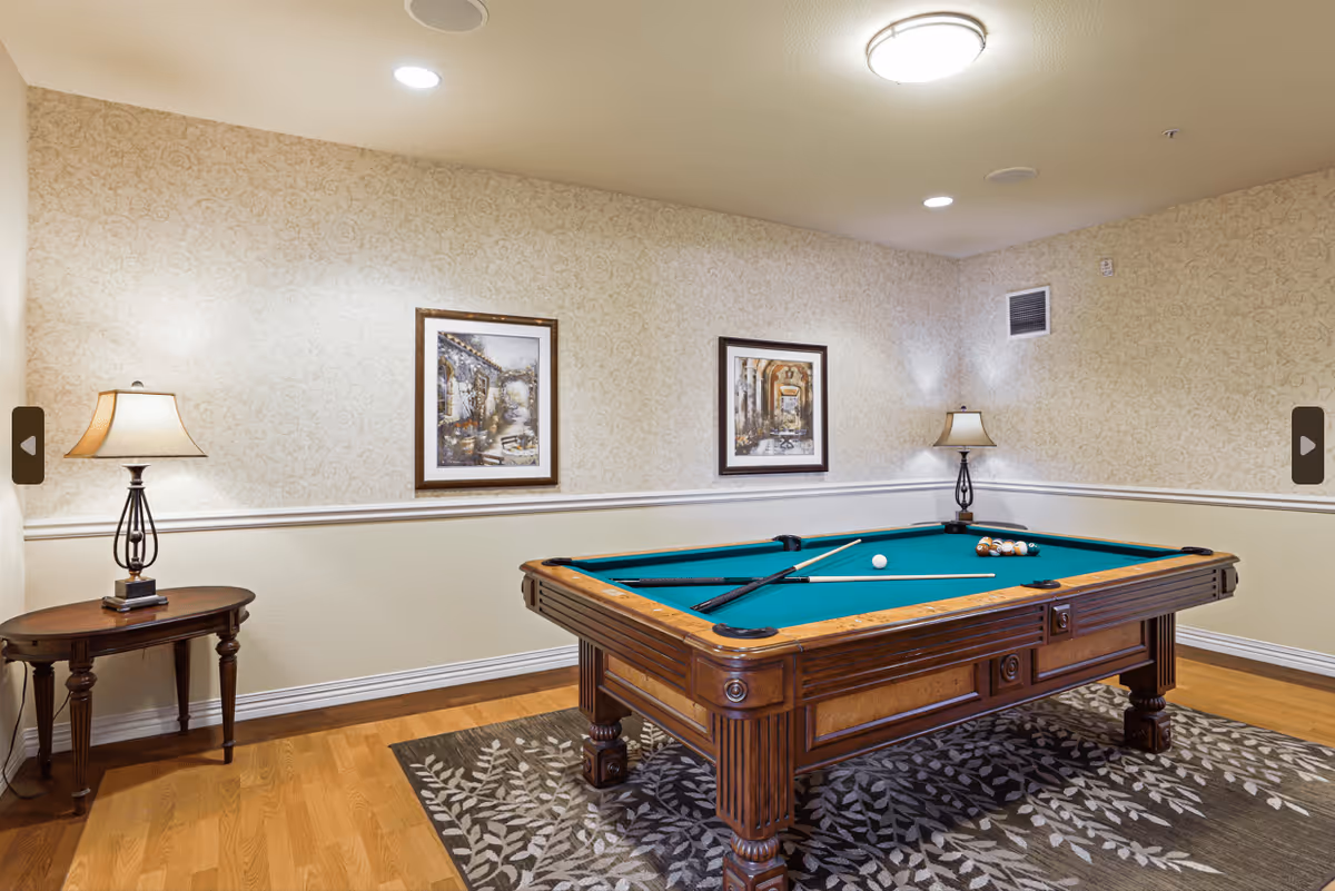 A well-lit room with a wooden pool table covered with a green felt surface. Two pool cues and a rack of billiard balls are on the table. The room has light-colored wallpaper with a subtle pattern, two framed pictures on the wall, two table lamps on small wooden tables, and a patterned area rug under the pool table.