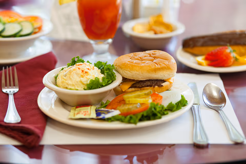 A plate with a cheeseburger, a bowl of potato salad garnished with lettuce, and a side of sliced tomato, pickles, and onion on a table set with a fork, knife, and spoon. In the background, there is a glass of iced tea and other plates with food.