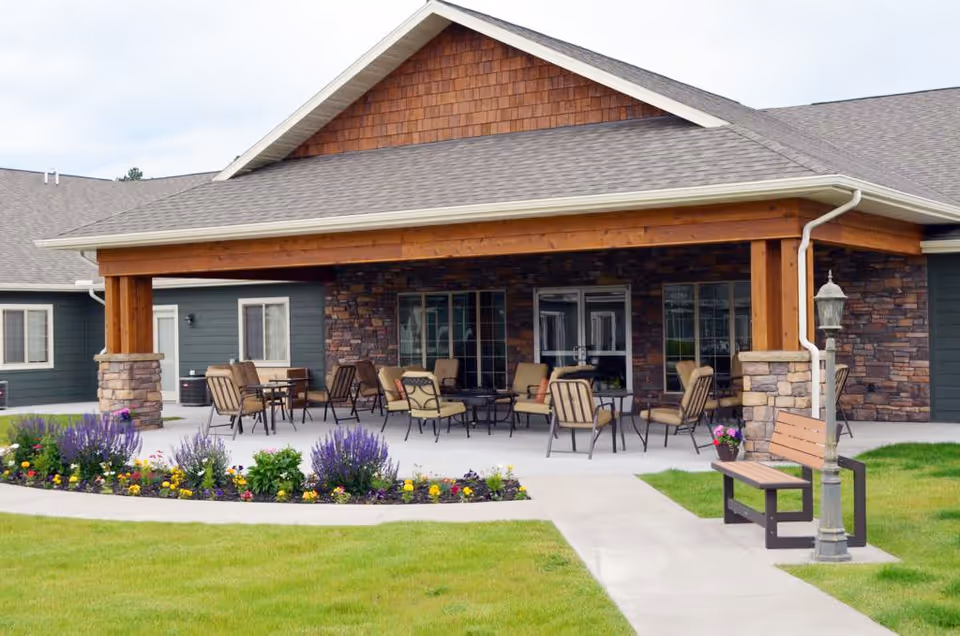 Covered outdoor patio at a senior living building with chairs and tables, a flower bed, and a bench in front.
