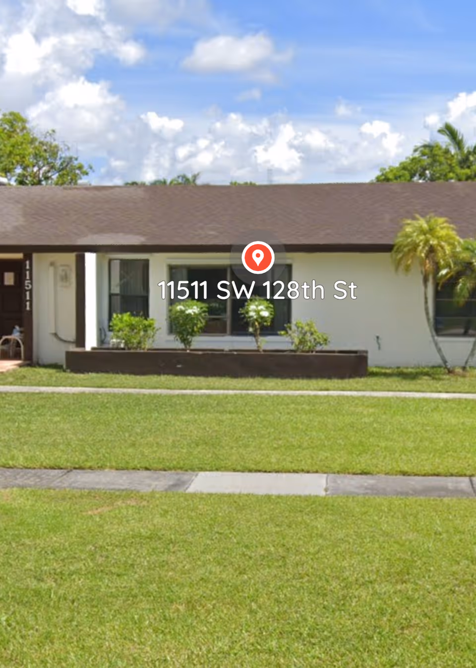 Single-story building with a brown roof and white walls, featuring a small garden bed with green shrubs in front and a palm tree on the right side. The building has windows and a door with the number 11511 displayed vertically. The sky is partly cloudy with blue patches.