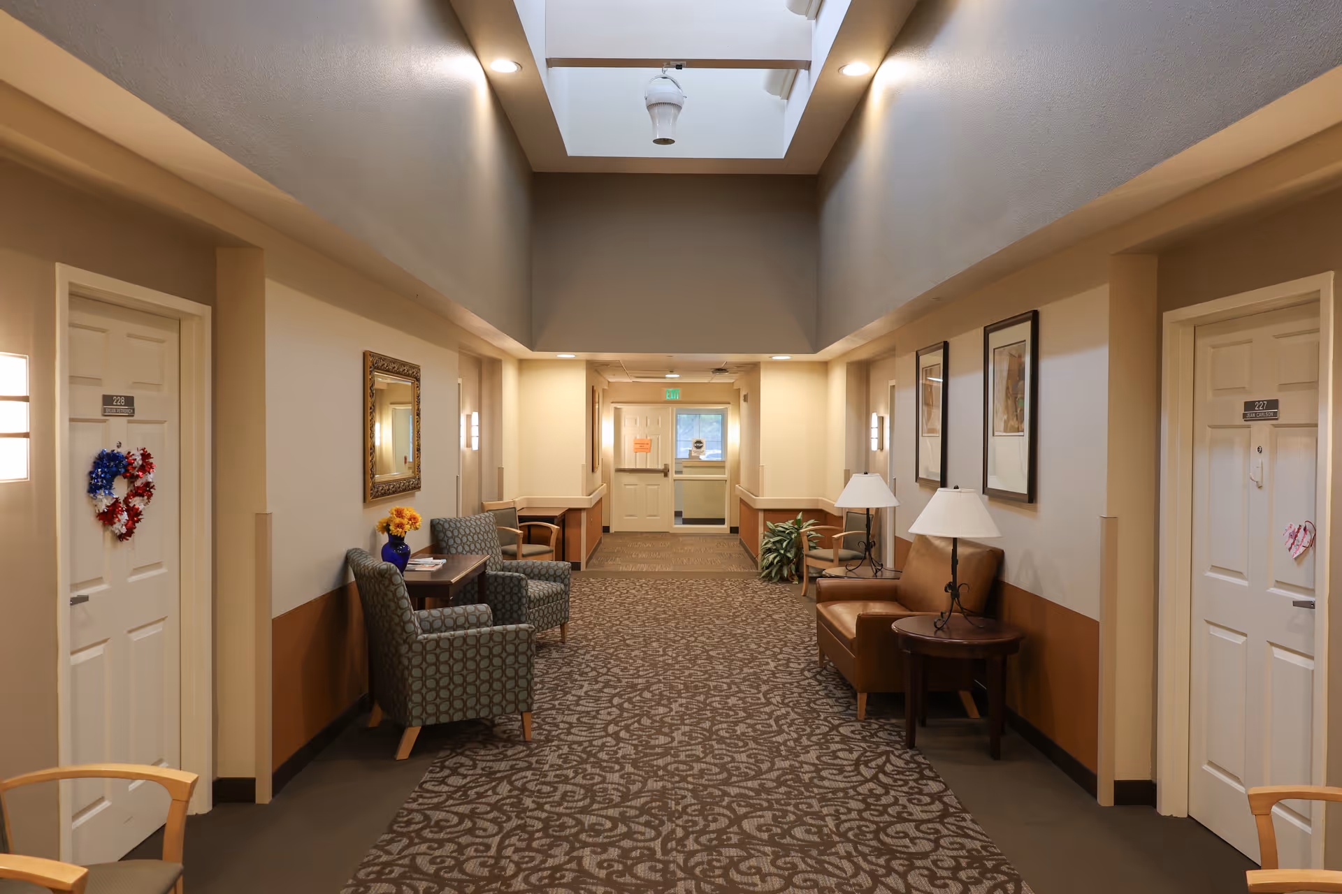 Well-lit interior hallway of a senior living facility with seating areas, room doors, and a skylight.