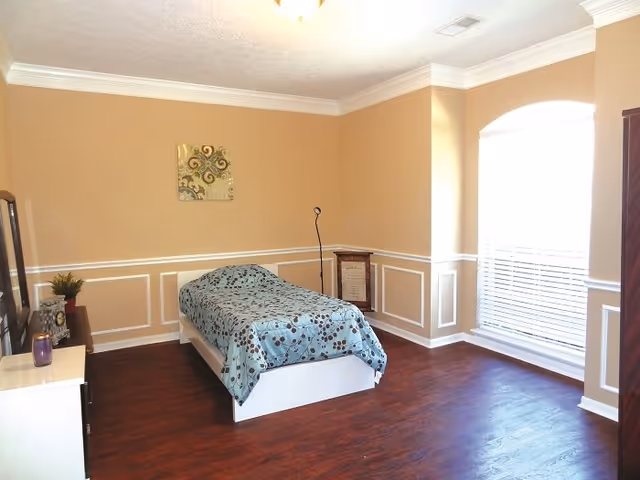 A bright furnished bedroom with a single bed covered by a patterned blue bedspread, a dresser and floor lamp against wainscoted beige walls and a large arched window with blinds.