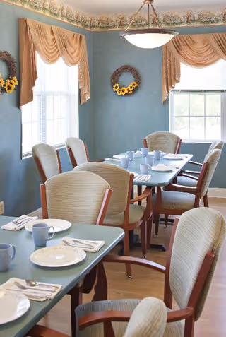 A dining room with multiple tables set with white plates, blue mugs, and silverware. The room has blue walls with floral wallpaper border near the ceiling, two windows with beige draped curtains, and several cushioned chairs with wooden armrests around the tables.