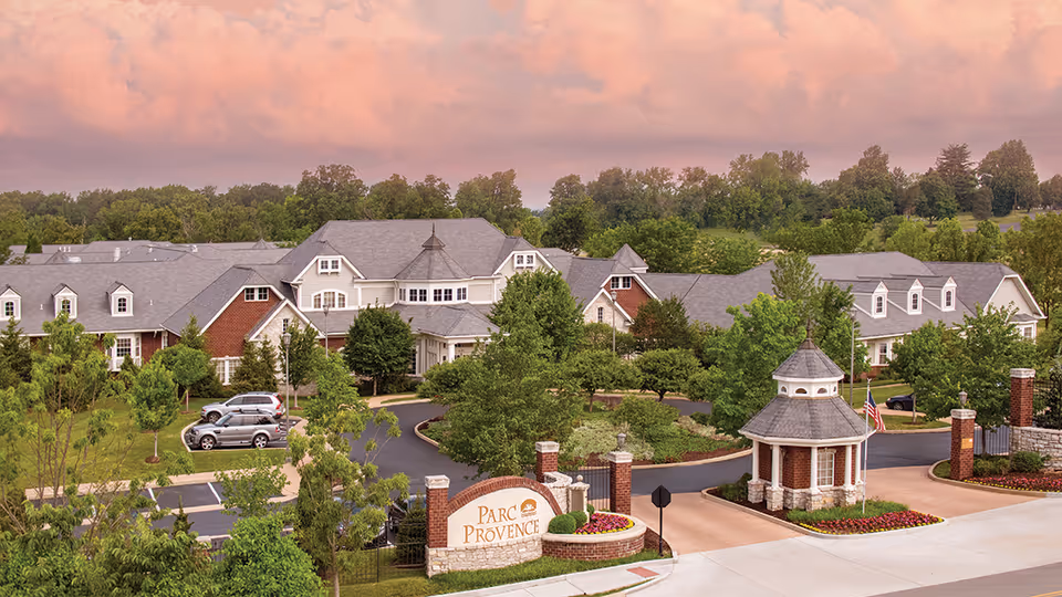 View of the Parc Provence senior living facility entrance with a circular driveway, landscaped greenery, and multiple connected buildings under a pinkish sky at sunset.
