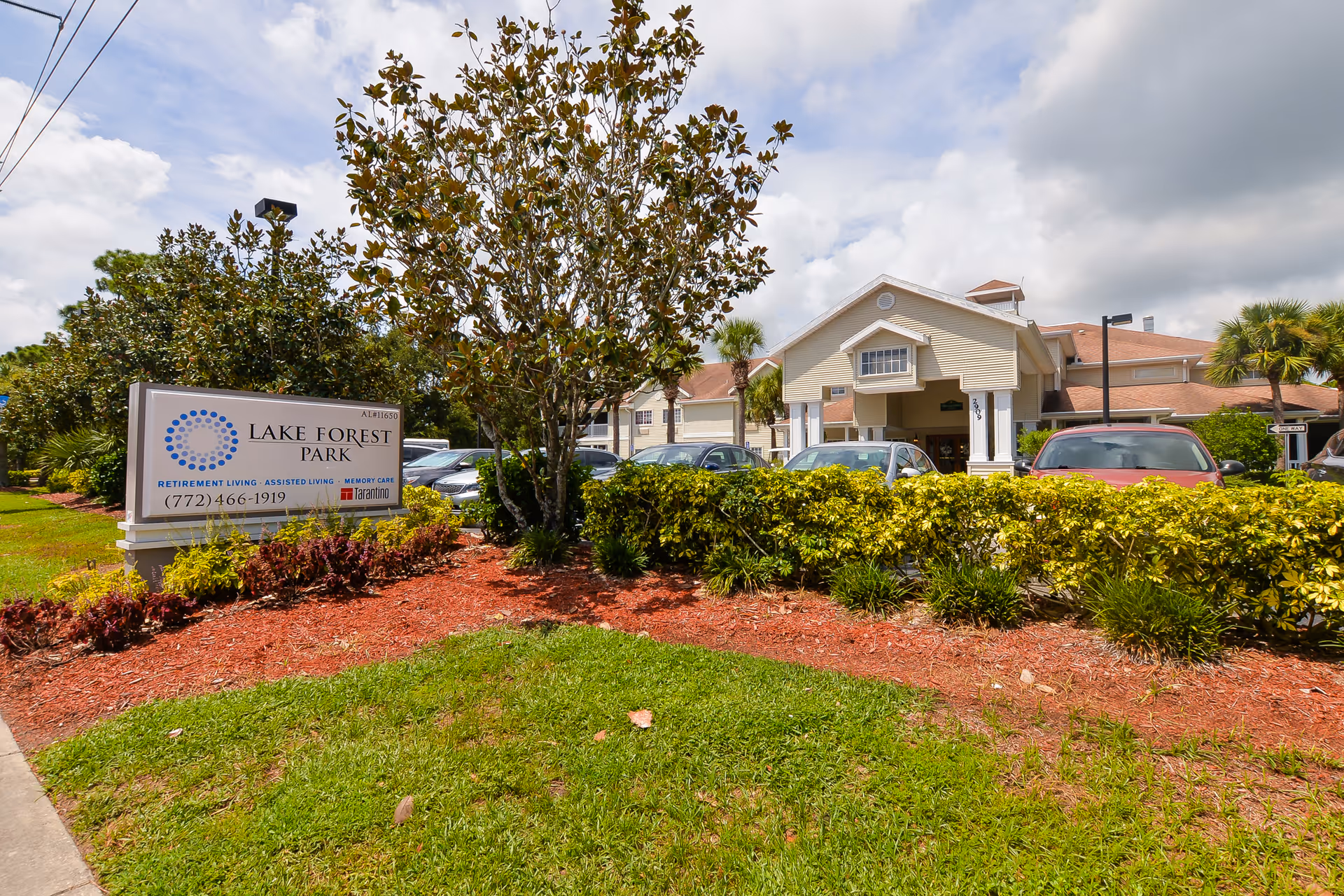 Front exterior of the Lake Forest Park senior living facility showing the entrance, parking lot, landscaping, and a freestanding sign.