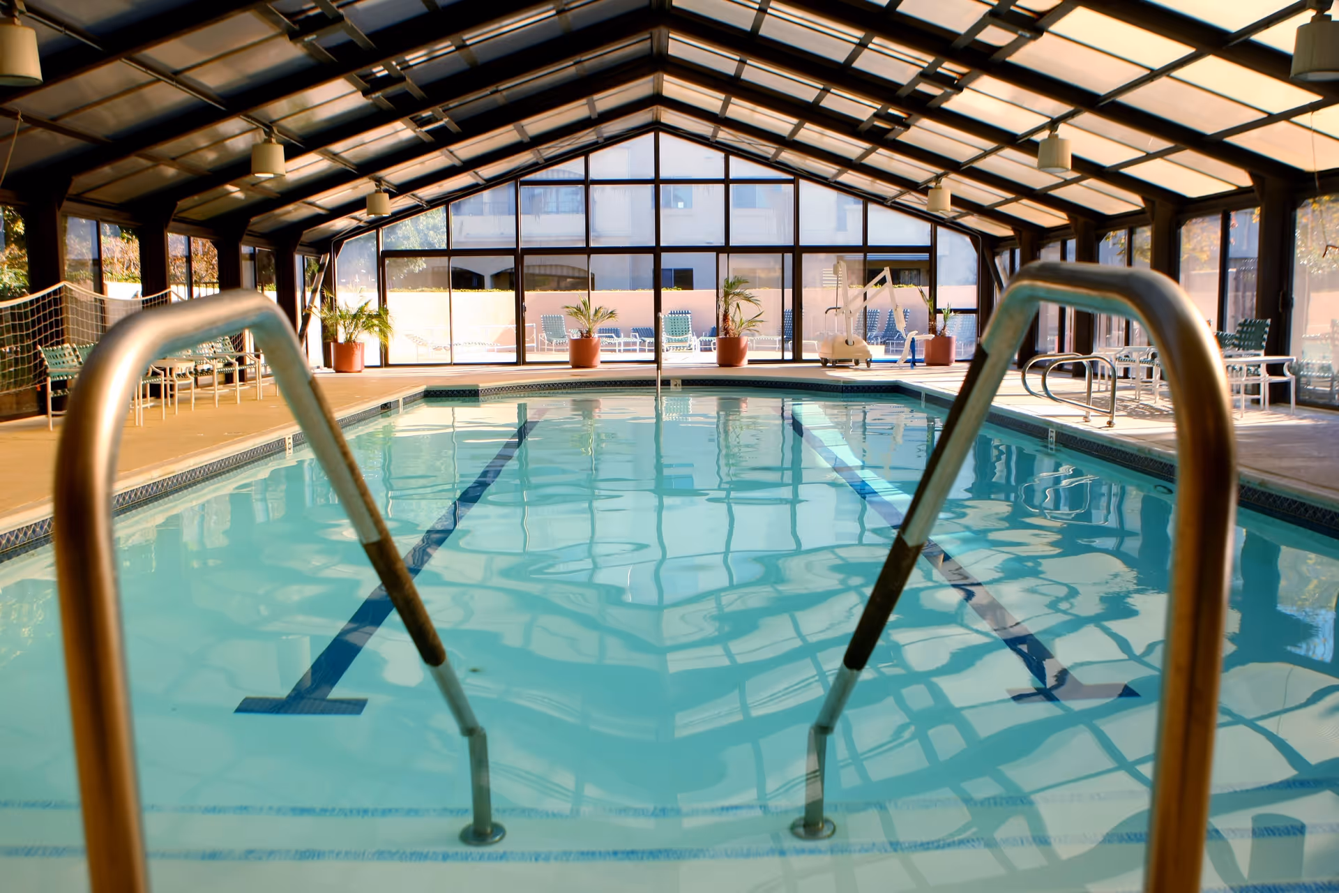 Indoor swimming pool with clear water, metal handrails in the foreground, surrounded by a glass enclosure with a slanted roof. There are potted plants and lounge chairs around the pool area.