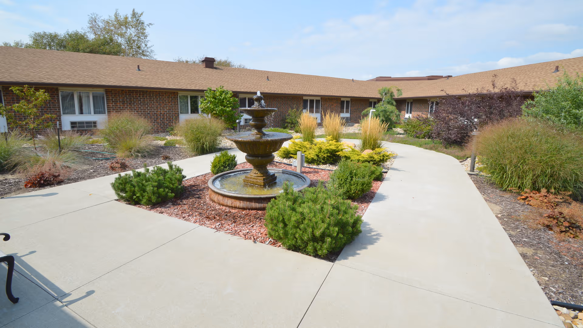 Outdoor courtyard area of a senior living facility with a central three-tiered water fountain surrounded by landscaped bushes and plants. The courtyard is paved with concrete walkways and bordered by a single-story brick building with windows and air conditioning units.