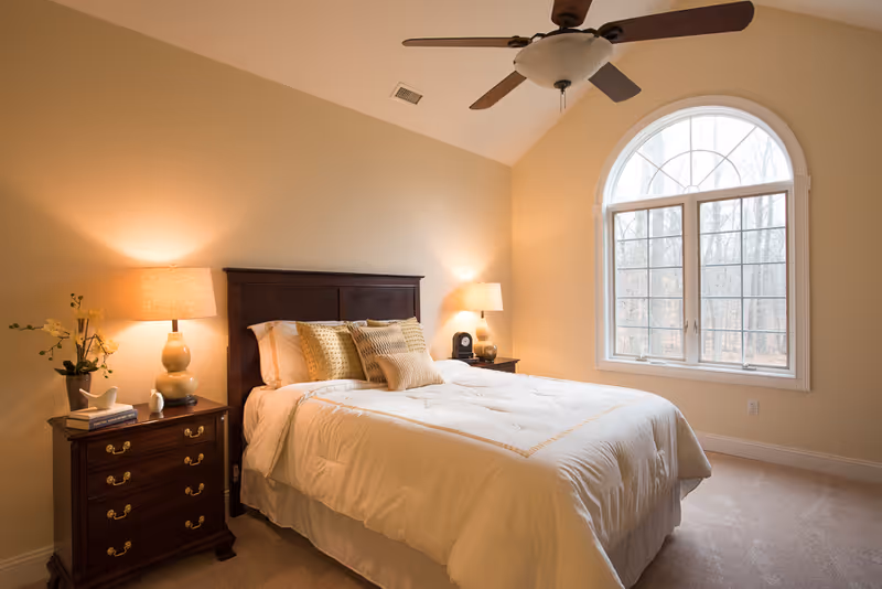 A cozy bedroom with a large bed covered in white bedding and multiple pillows. There are two wooden nightstands on either side of the bed, each with a lamp. A large arched window lets in natural light, and a ceiling fan is mounted above the bed. The walls are painted a soft beige color.