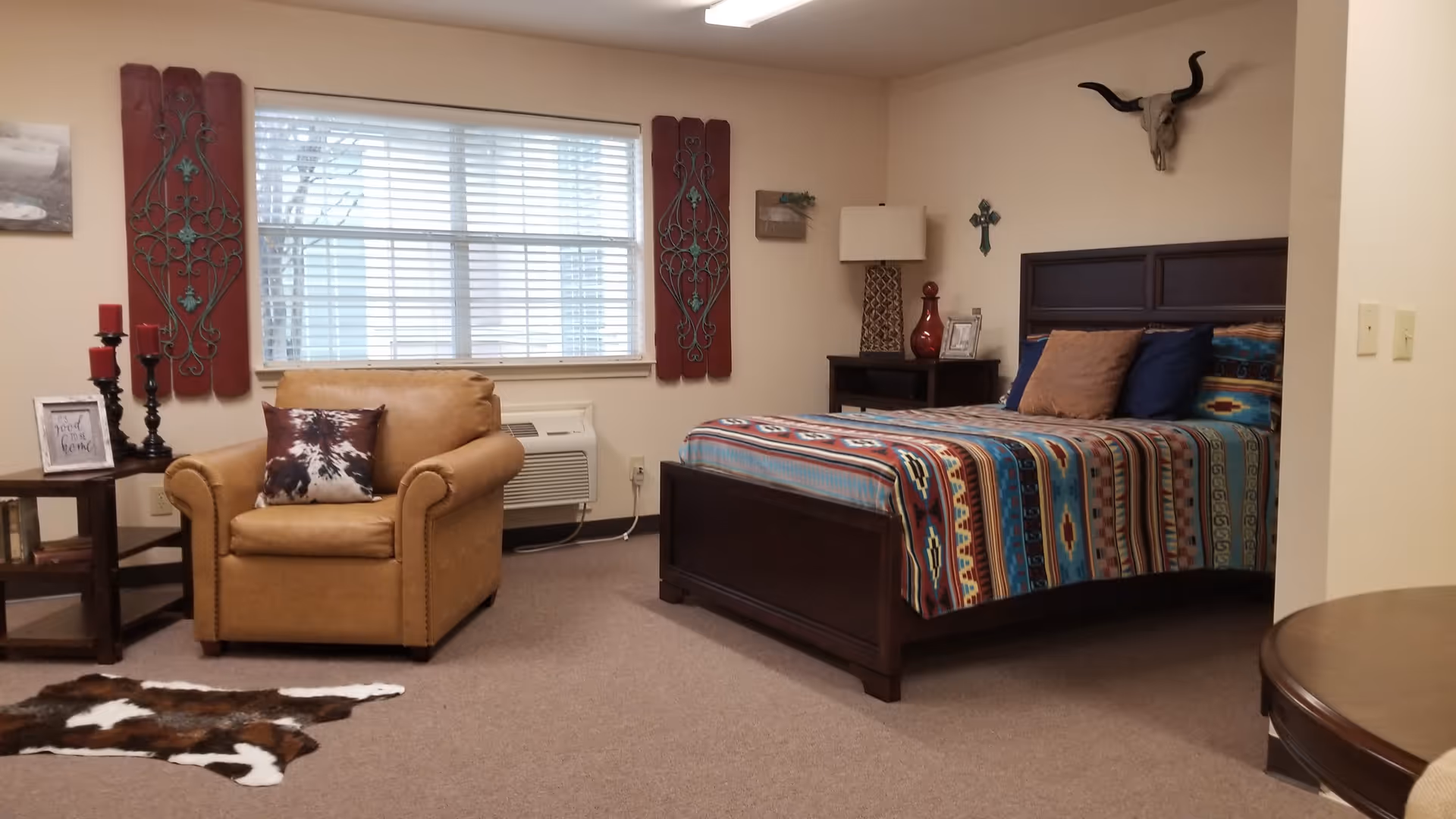 A cozy bedroom in an assisted living facility featuring a bed with a colorful southwestern patterned bedspread, a brown armchair with a decorative pillow, a side table with red candles and a framed sign, a window with blinds, and wall decorations including a skull with horns and a cross.