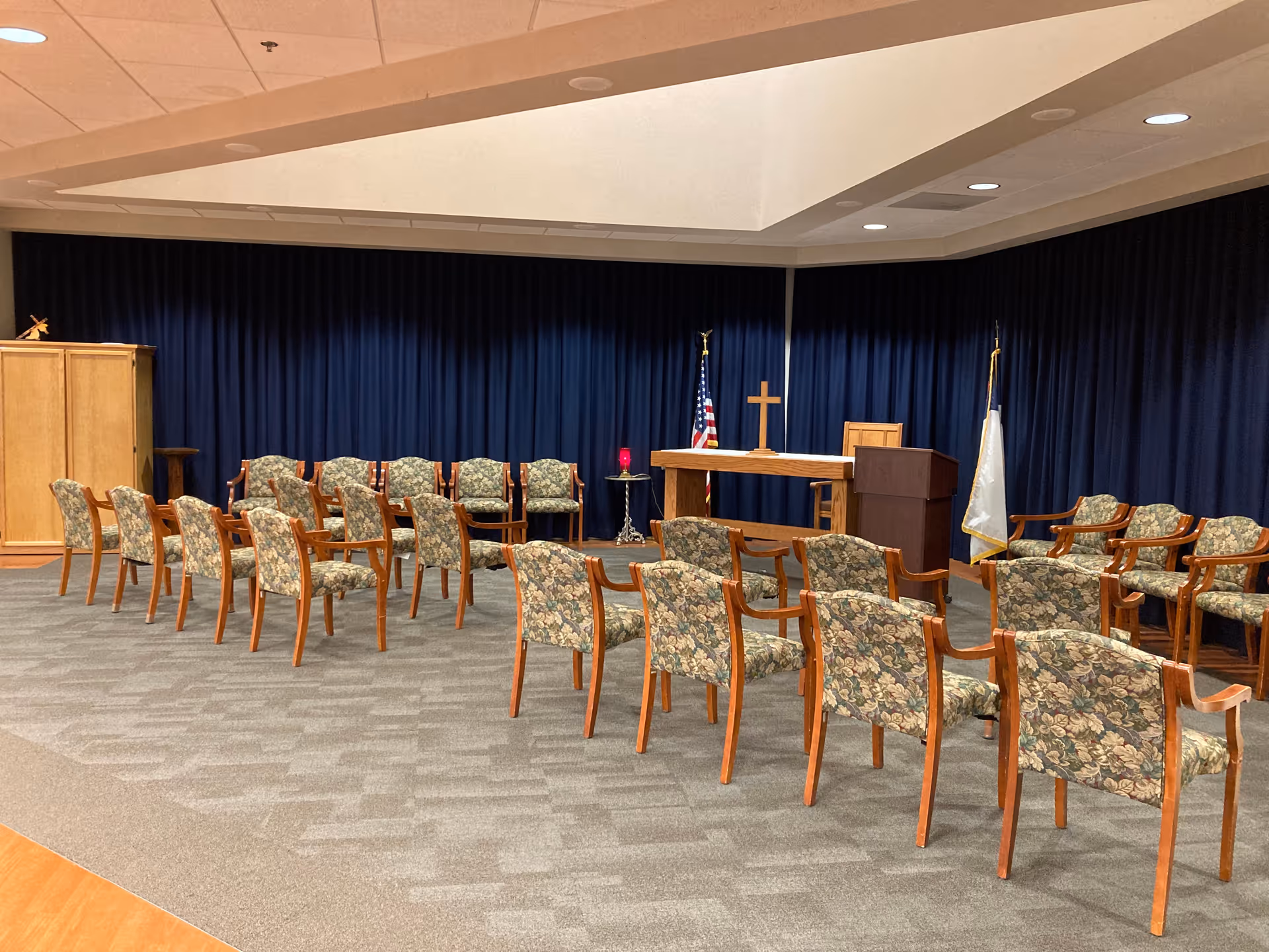 A chapel room with rows of floral upholstered wooden chairs arranged facing a wooden altar with a cross, an American flag, and another flag. The room has a blue curtain backdrop and a carpeted floor.