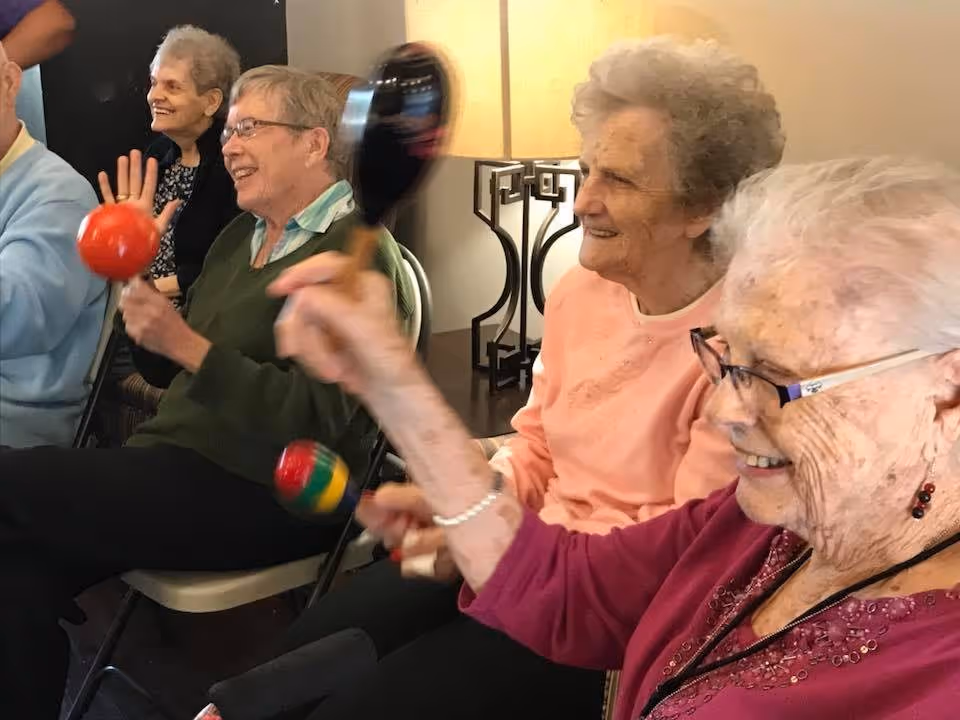 A group of elderly women sitting in chairs indoors, smiling and shaking colorful maracas, participating in a musical activity. A lamp and a table are visible in the background.