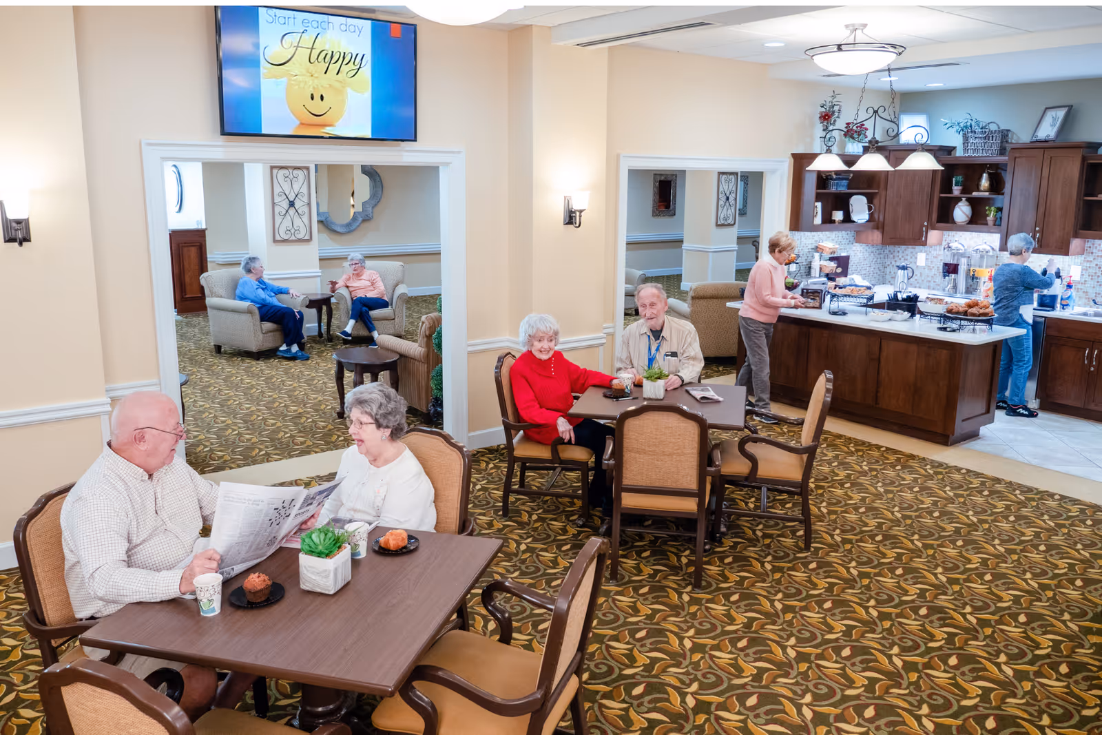 A bright and welcoming common area in a senior living facility with several elderly residents. Two couples are seated at separate tables, engaging in conversation and enjoying snacks. In the background, two women are preparing food and drinks in a kitchen area with wooden cabinets and a countertop filled with various items. The room has patterned carpet, beige walls, and soft lighting. A TV mounted on the wall displays a cheerful message: 'Start each day Happy.'