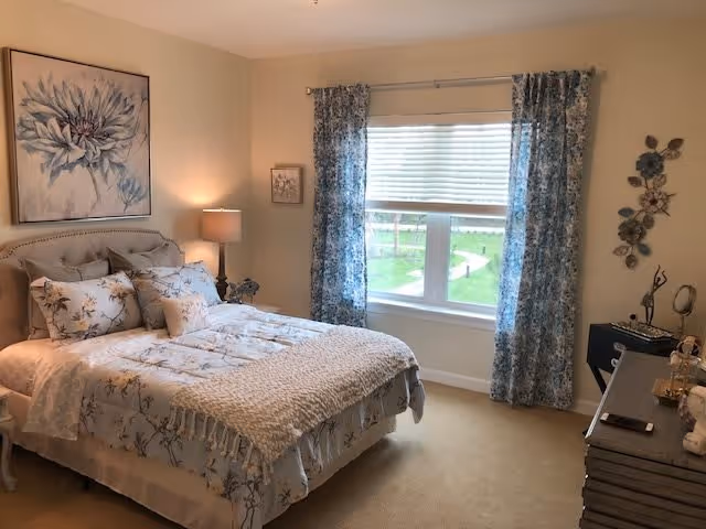 A cozy bedroom with a bed dressed in floral bedding and a knitted throw blanket. There is a large window with blue patterned curtains letting in natural light. The room features a bedside table with a lamp, a dresser with decorative items, and wall art including a large flower painting and a metal floral wall decoration.