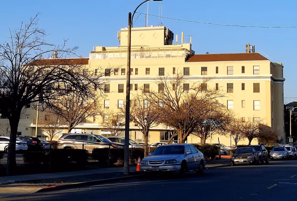 Exterior view of a multi-story beige hospital building with several windows, a parking lot with cars in front, leafless trees, and a street with parked cars under a clear blue sky.
