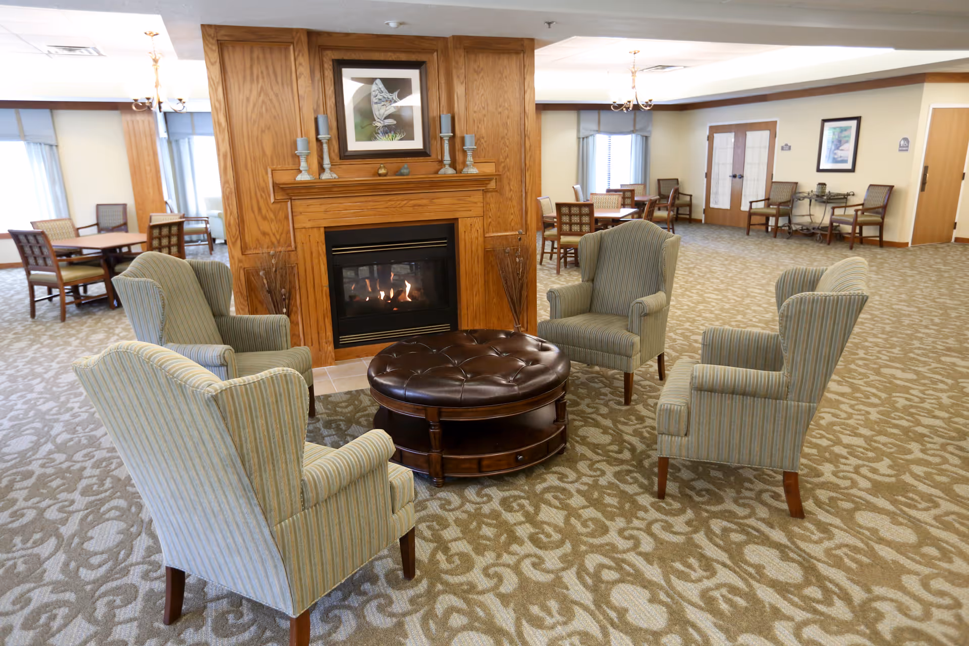A senior living common room with a fireplace, four upholstered armchairs arranged around a round leather ottoman and dining tables in the background.