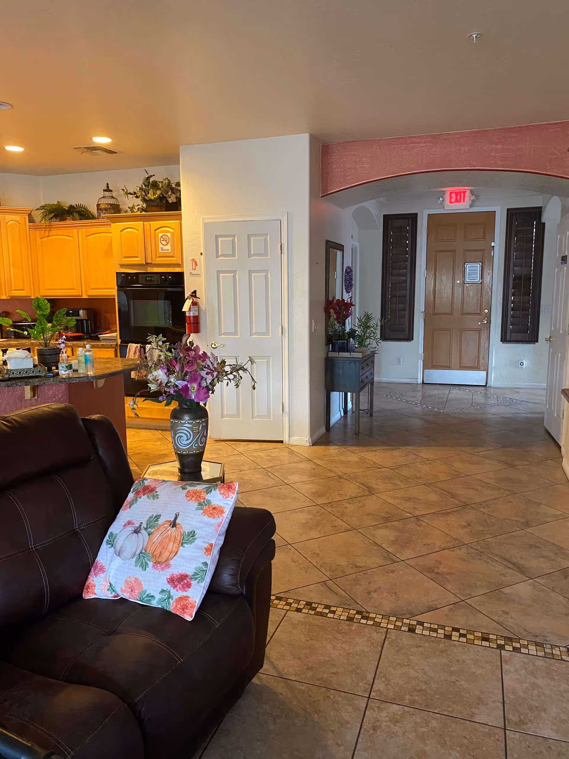 Interior view of a living area in an assisted living home showing a brown leather couch with a decorative pillow featuring pumpkins and flowers. A vase with purple flowers is on a small glass table next to the couch. In the background, there is a kitchen with wooden cabinets, a built-in oven, and various plants and items on the counter. The floor is tiled, and there is an arched doorway leading to a hallway with a wooden door and an exit sign above it.