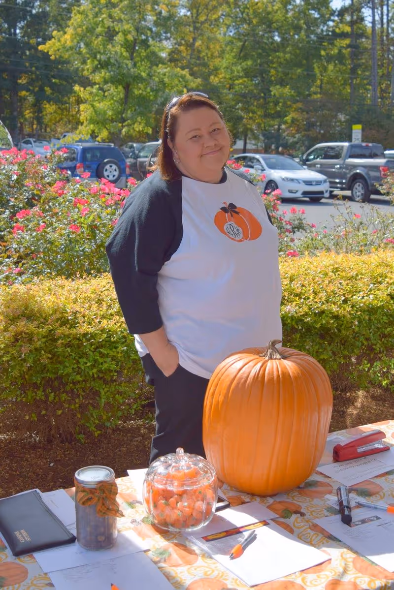 A woman standing outdoors behind a table decorated with a large pumpkin, a glass jar filled with small orange pumpkins, and various papers and pens. She is wearing a white and black shirt with a pumpkin design and smiling. Behind her are bushes, flowers, parked cars, and trees.