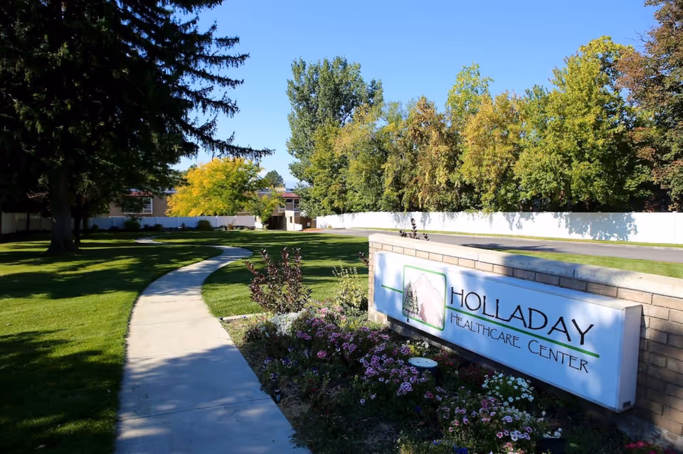A sunny outdoor scene at Holladay Healthcare Center featuring a curved concrete walkway through a well-maintained green lawn with trees and flower beds. A brick sign with the facility's name is prominently displayed in the foreground.
