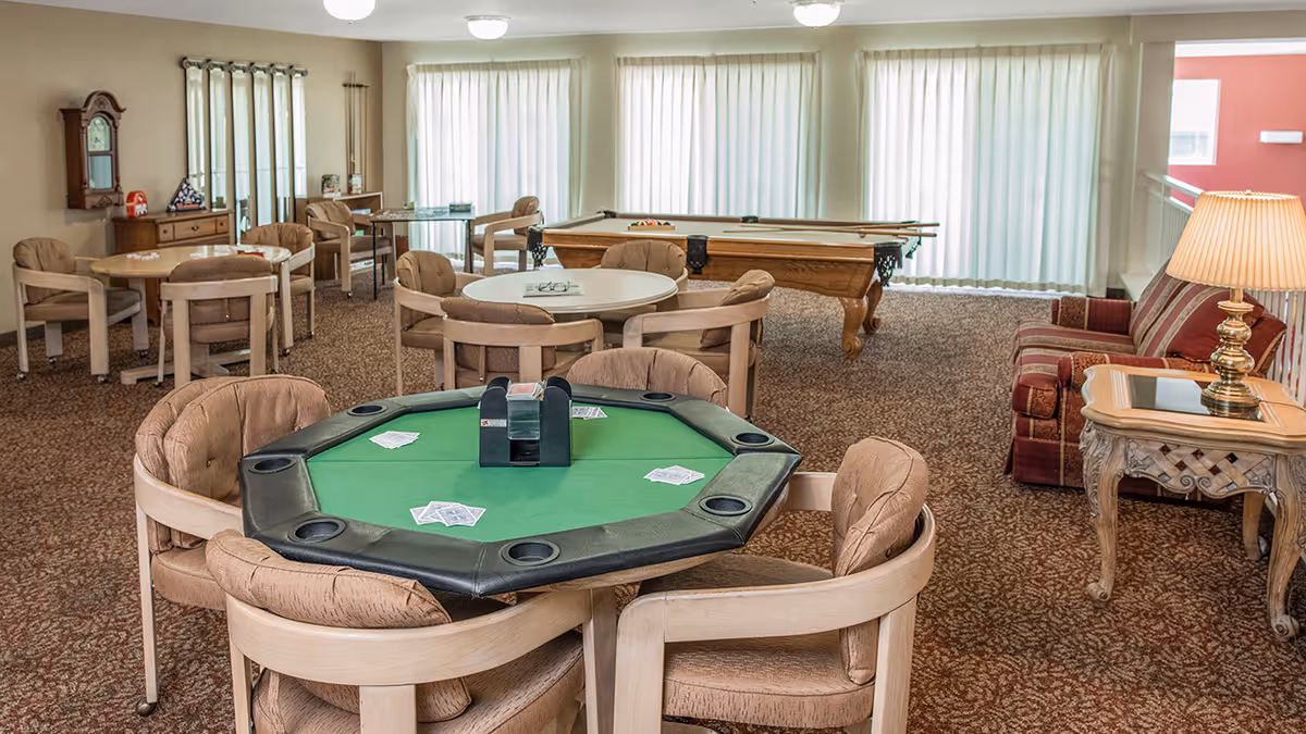 A senior living facility game room with a green felt poker table surrounded by cushioned chairs, several round tables with chairs, a pool table in the background, a striped couch with a side table and lamp, and large windows with vertical blinds letting in natural light.