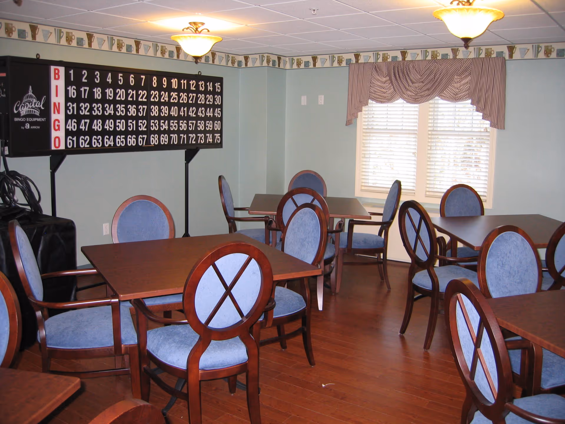 A room with several wooden tables and blue cushioned chairs arranged around them. On the wall, there is a large bingo board with numbers and the word BINGO vertically displayed. The room has wooden flooring, light green walls, two ceiling lights, and a window with striped valance curtains.