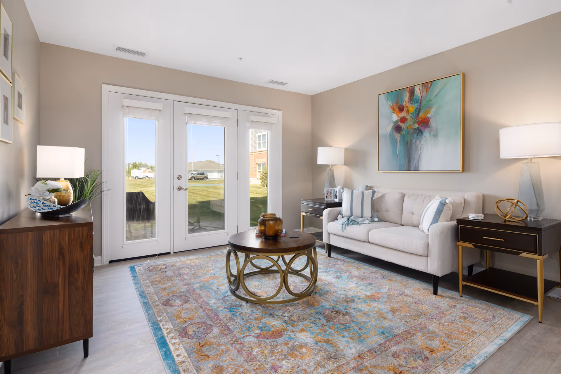 A bright and modern living room featuring a beige sofa with striped pillows, two side tables with lamps, a round coffee table with decorative items, a colorful area rug, and large glass doors leading outside. The walls are painted light beige and adorned with framed artwork.