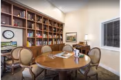 A cozy interior room with a round wooden table surrounded by six upholstered chairs. Behind the table is a large wooden bookshelf filled with books and decorative items. A clock and framed pictures are on the walls, and a piano with a lamp is positioned near a window with blinds.