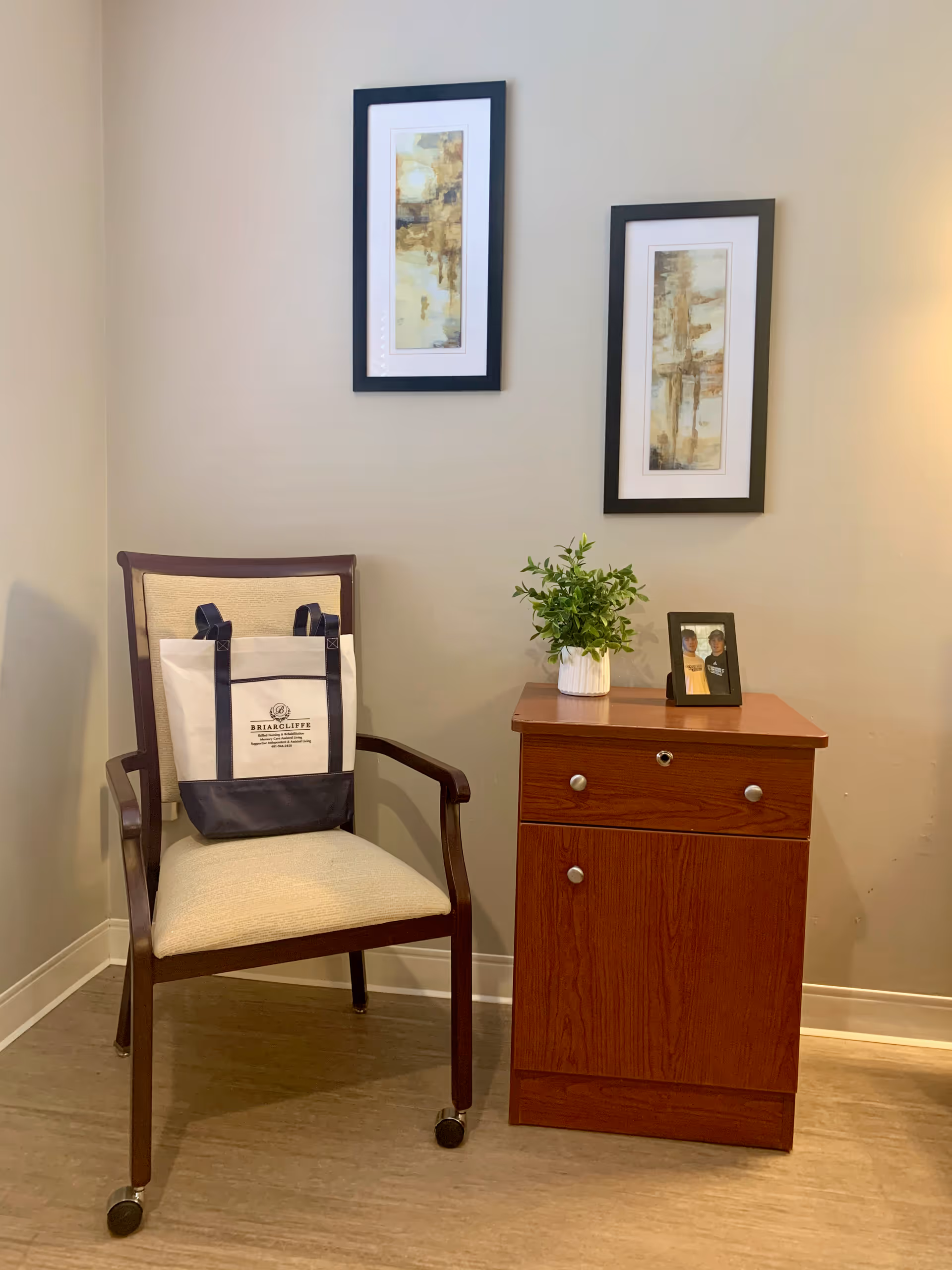 A corner of a room with a wooden chair that has a beige cushion and a tote bag hanging on it. Next to the chair is a wooden cabinet with a small green plant in a white pot and a framed photo on top. Two vertical abstract paintings in black frames hang on the beige wall above.