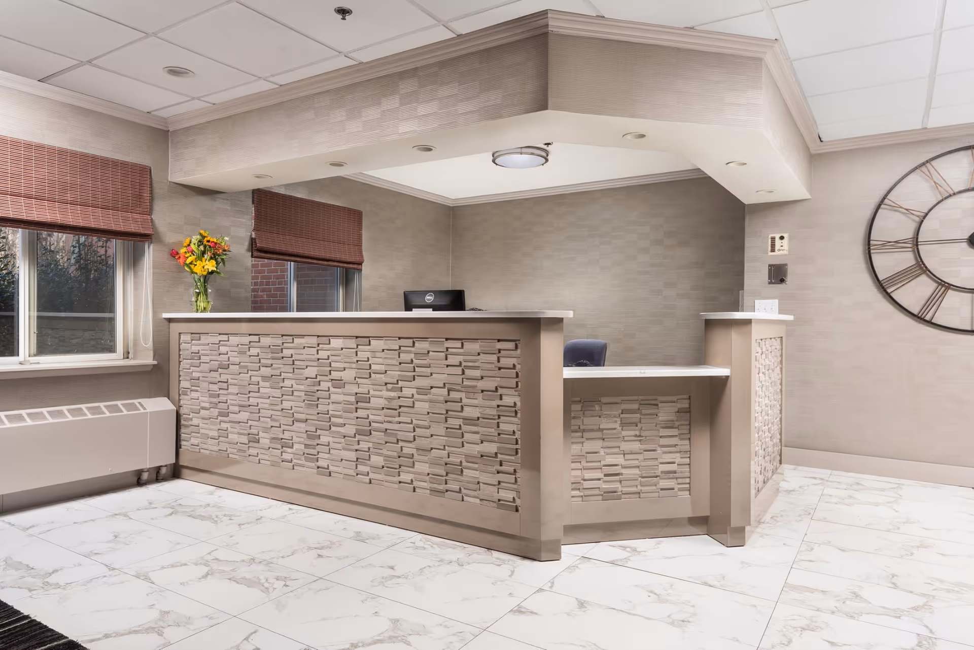 Reception desk in a neutral-toned lobby with marble tile floor, a vase of flowers, and a large wall clock.