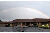 Exterior view of a single-story brick building with a covered entrance and a wet parking lot in front. A rainbow arcs across the cloudy sky above the building.