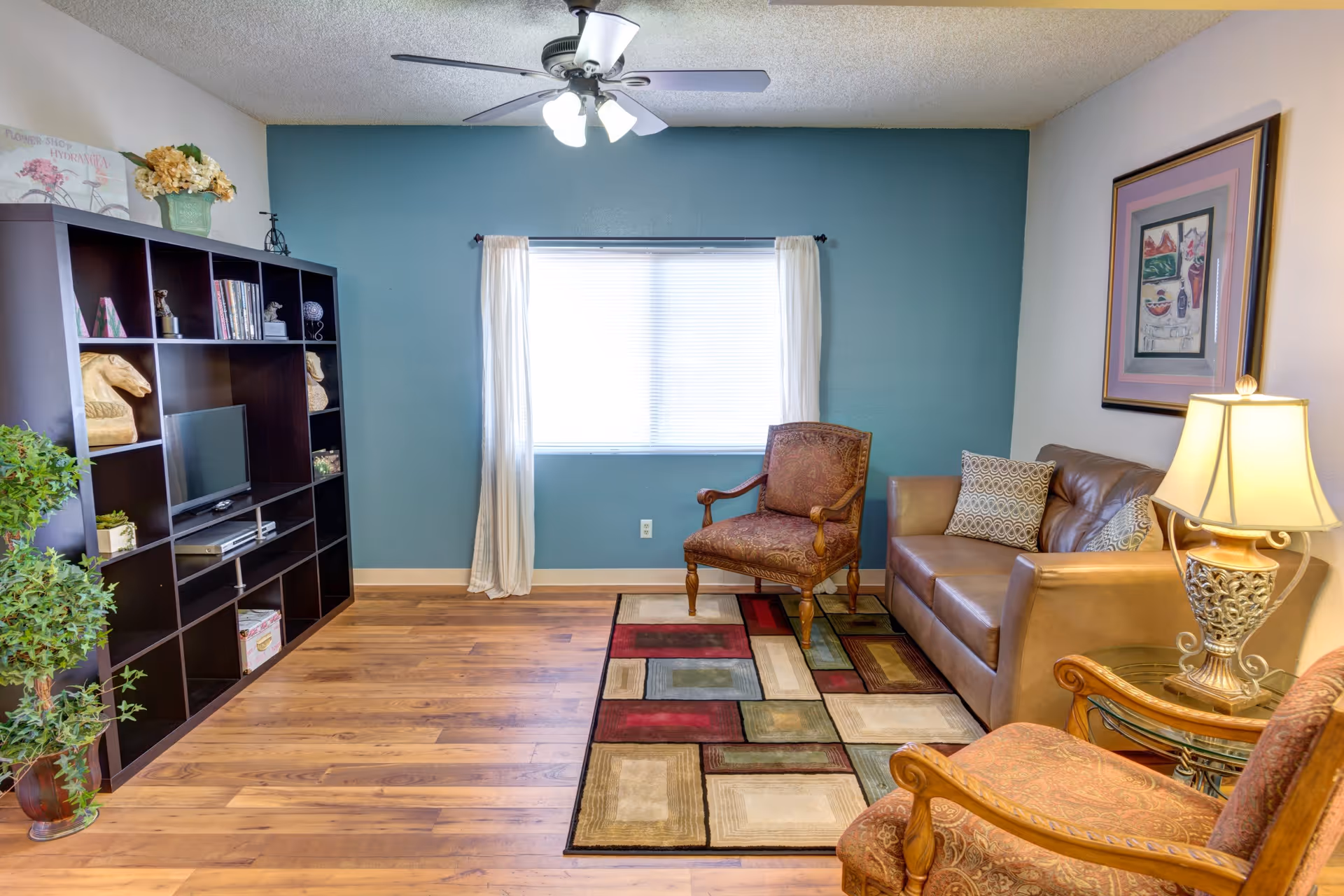 A cozy living room with a wooden floor, a colorful geometric patterned rug, a brown leather sofa with patterned cushions, two upholstered armchairs with wooden frames, a glass side table with an ornate lamp, a large dark wood shelving unit with decorative items and a small TV, a potted plant, and a window with white curtains on a teal accent wall.