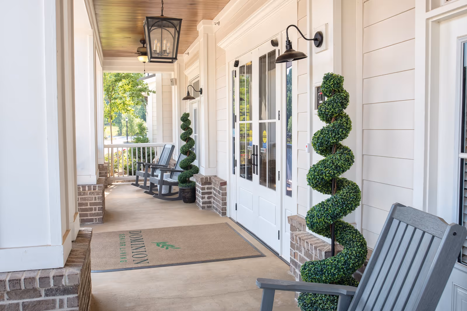 Covered porch entrance with rocking chairs, spiral topiary plants, and double glass doors leading into a senior living facility.