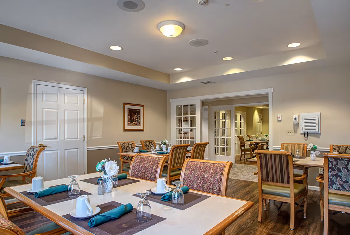 A dining room in a senior living facility with several tables set with placemats, cups, glasses, and napkins. The room has wooden chairs with patterned upholstery, beige walls, and a ceiling with recessed lighting. There is a door on the left wall and a set of glass-paneled double doors leading to another room with more tables and chairs.