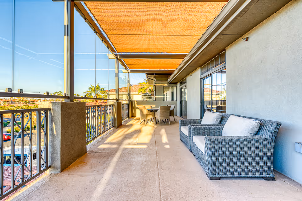 A spacious outdoor balcony area with a wicker loveseat with cushions on the right and a round table with four wicker chairs further down. The balcony has a railing with decorative metalwork and a partial glass windbreak. An orange shade canopy covers the top, and the view includes rooftops and palm trees under a clear blue sky.