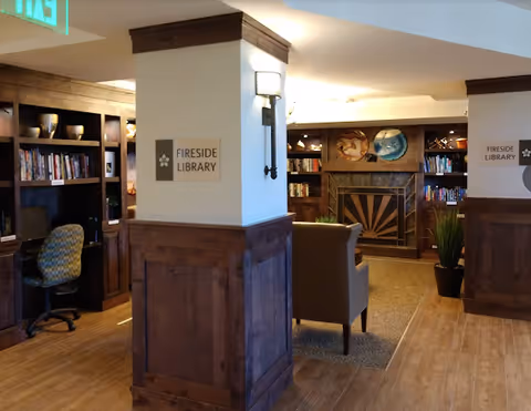 Interior view of a cozy library room named Fireside Library with wooden bookshelves filled with books, a computer workstation with a chair, a fireplace with decorative artwork above it, and comfortable seating including an armchair. The room has warm lighting and wood paneling on the walls.