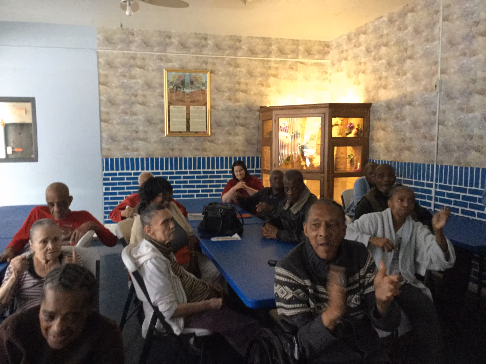 A group of elderly residents seated around blue tables in a communal room at a senior living facility.