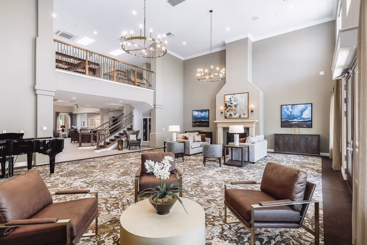Spacious senior living facility common area with high ceilings, two large chandeliers, a grand piano, multiple seating arrangements including leather chairs and sofas, a fireplace with artwork above it, and large windows with curtains allowing natural light.