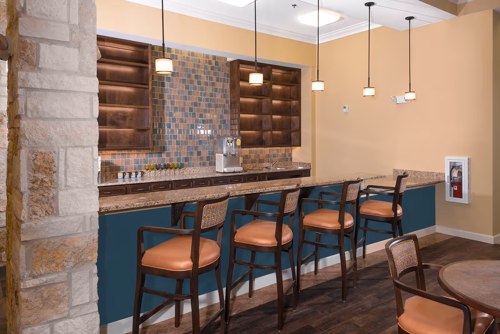 Interior view of a seating area with a long counter and five high chairs with brown cushions. Behind the counter are empty wooden shelves and a tiled backsplash with a beverage dispenser on the countertop. The walls are painted beige and there are four pendant lights hanging from the ceiling.