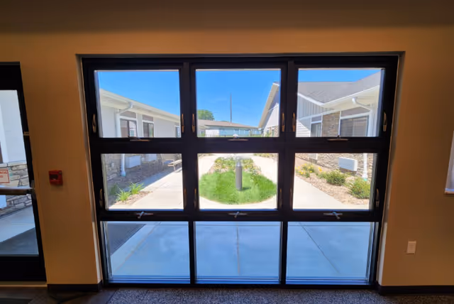 Interior view through a large multi-pane glass door looking out to a courtyard walkway and adjacent single-story memory care buildings.