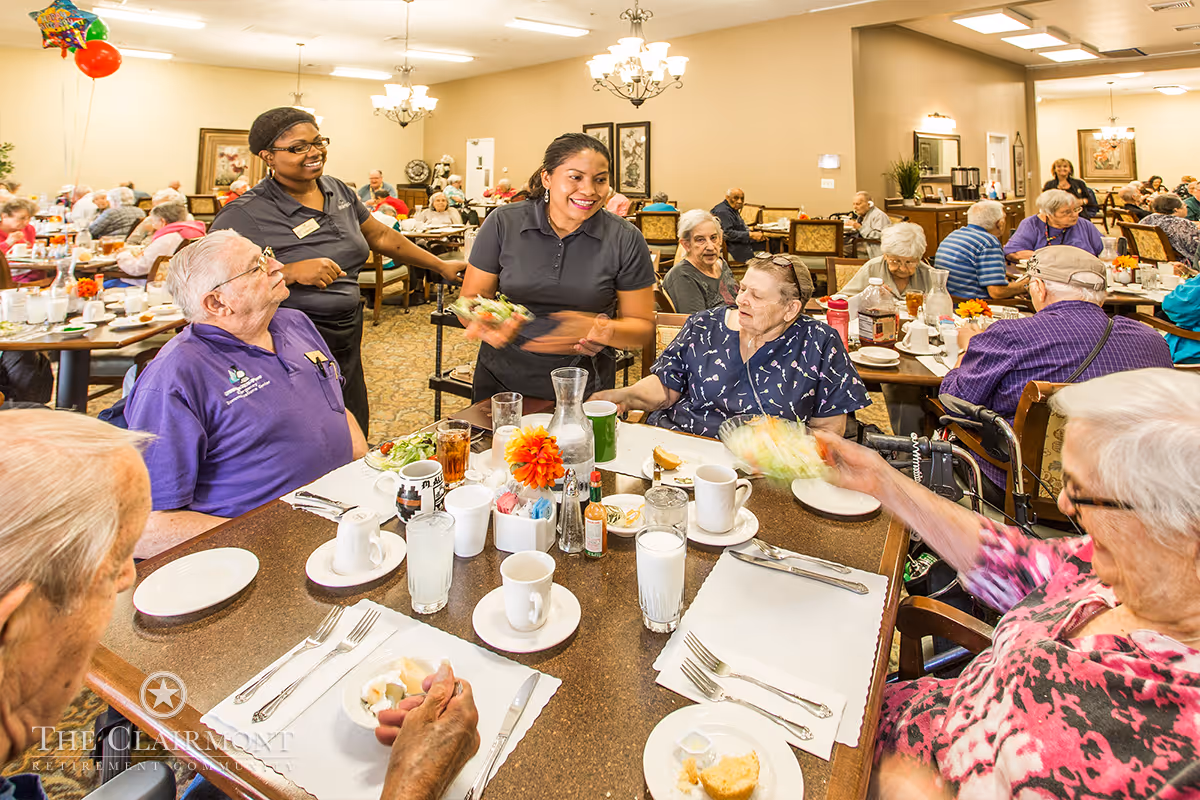 Staff serving meals to elderly residents seated around a large communal dining table in a busy retirement community dining room.