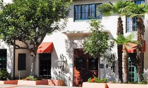 Facade of Villa Santa Barbara senior living facility with red doors, awnings, and palm trees.