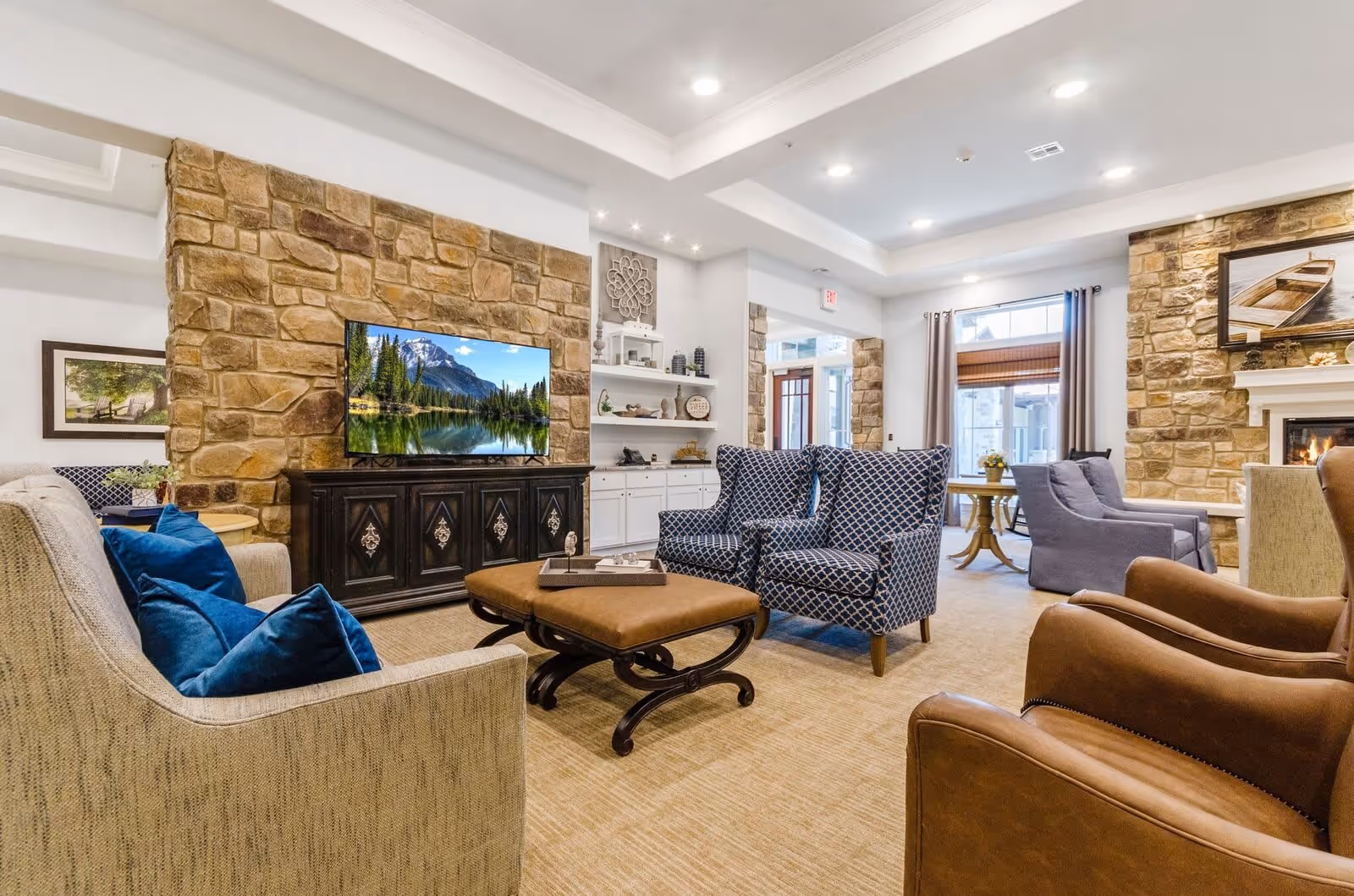 A cozy living room in a senior living facility featuring a stone accent wall with a mounted flat-screen TV displaying a mountain and lake scene. The room has various seating options including a beige sofa with blue pillows, two patterned blue armchairs, two brown leather chairs, and two gray armchairs near a fireplace with a boat painting above it. The space is well-lit with recessed ceiling lights and has a beige carpeted floor.